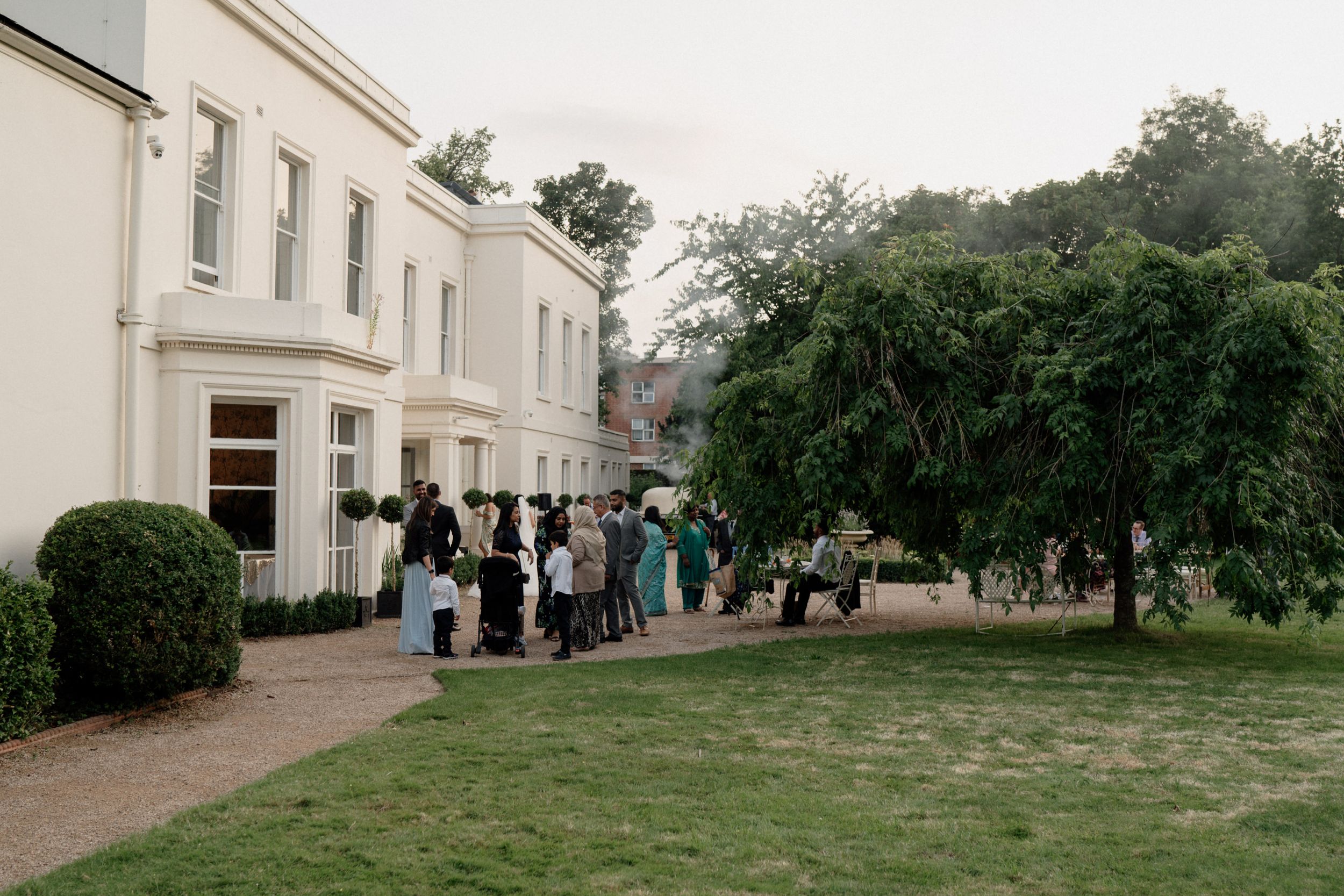 Guests gather outside an elegant white manor house of Morden Hall in London with manicured gardens and topiary bushes on a cloudy day.