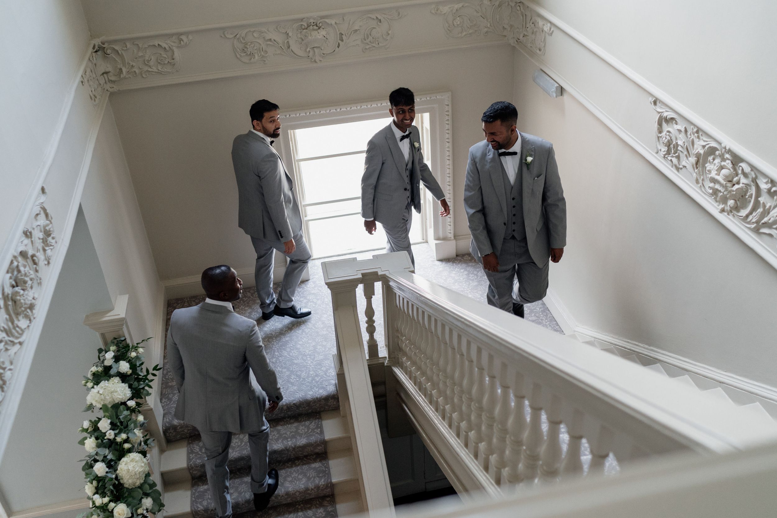 A group of groomsmen in matching gray suits descend an elegant white staircase decorated with floral arrangements.