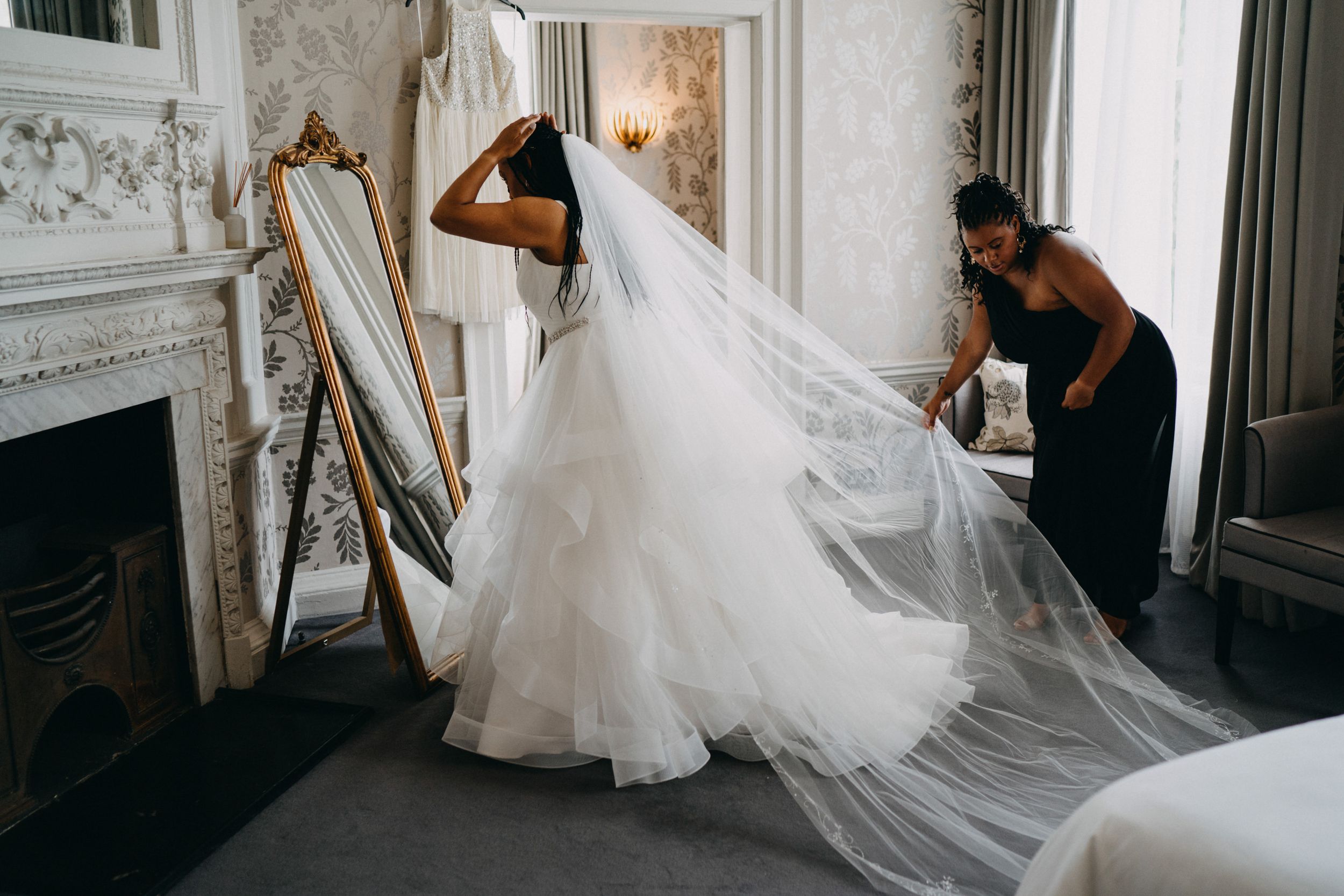 A flowing white wedding dress and veil are being arranged in an elegant room with ornate fireplace and mirror.
