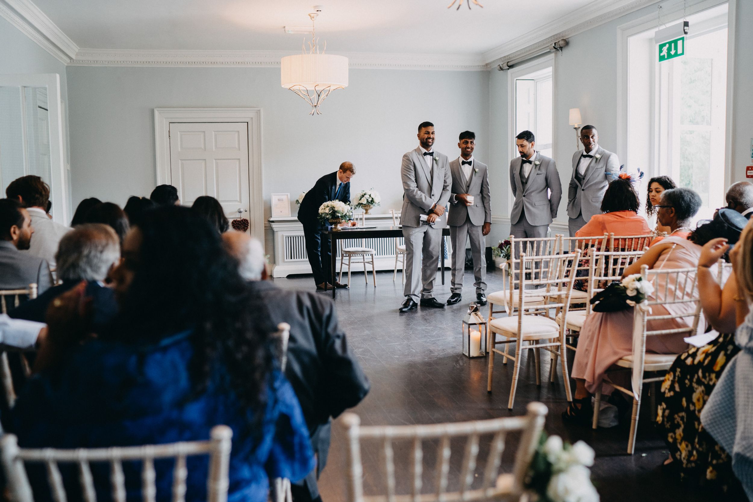 Wedding ceremony at Morden Hall guests sit in white chairs watching groomsmen at the altar in a bright modern venue.