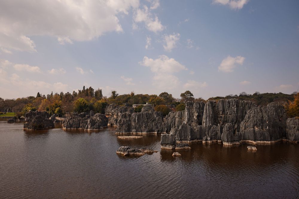 Stone Forest scenic area with dramatic limestone rock formations rising from calm lake waters under blue sky and clouds.