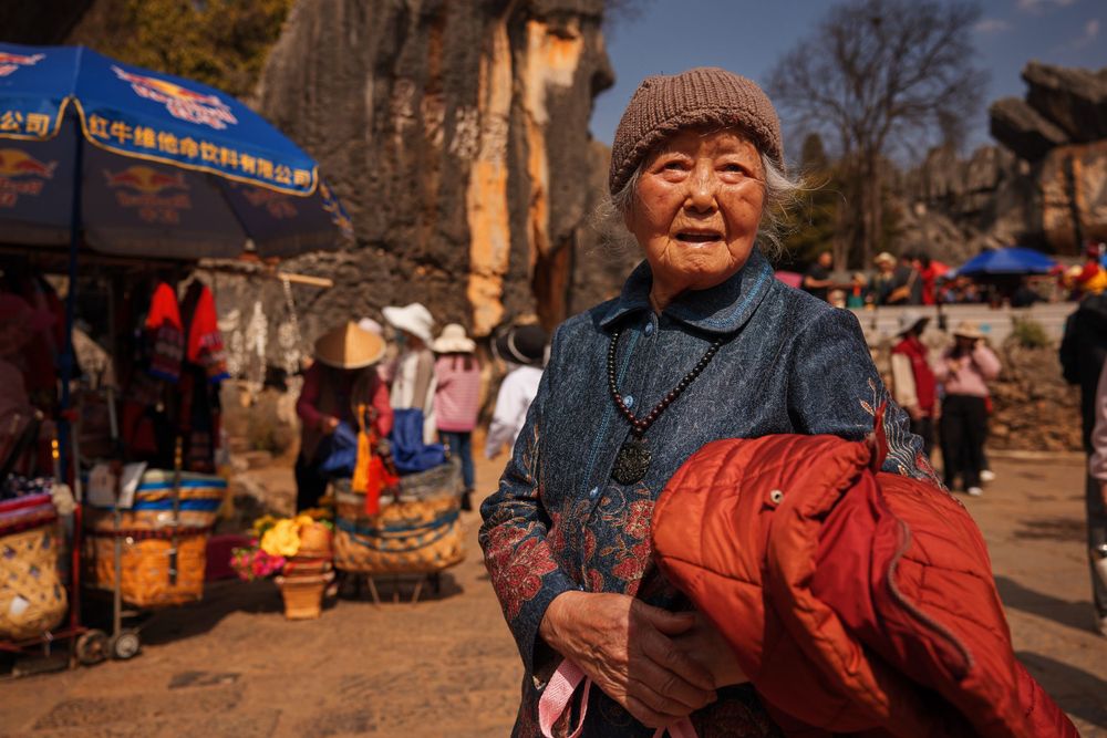 A person wearing a brown cap and blue sweater carries a red bundle at an outdoor market in autumn.