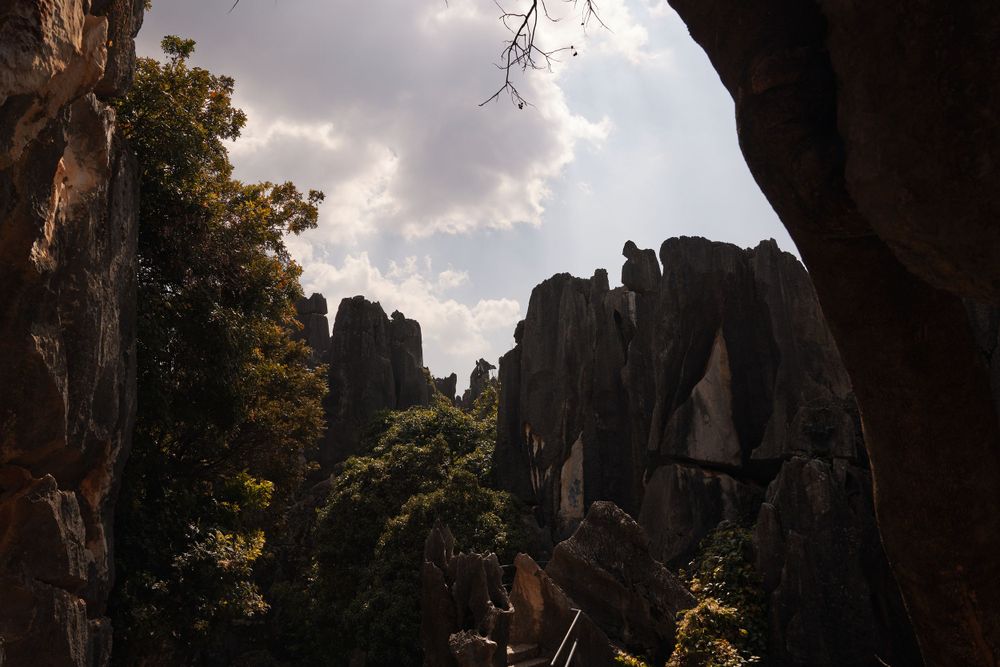 Dramatic rock formations silhouetted against cloudy sky with sunlight peeking through trees at sunset.