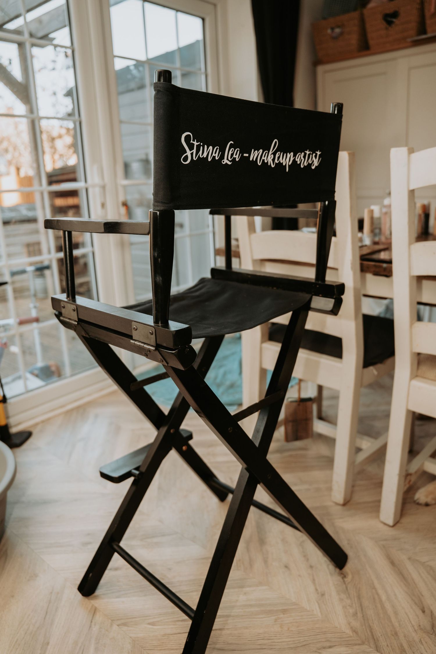 A black professional makeup artist chair with custom text on the backrest sits in a bright studio with herringbone flooring.
