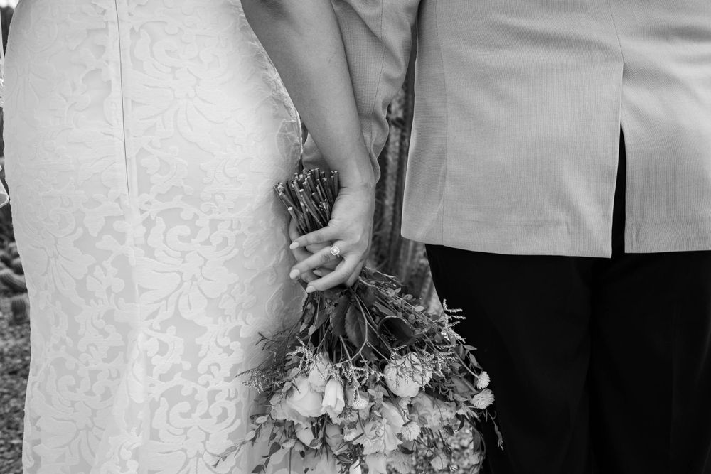 Black and white wedding photo detail showing bride in lace dress holding rustic floral bouquet next to groom's suit.