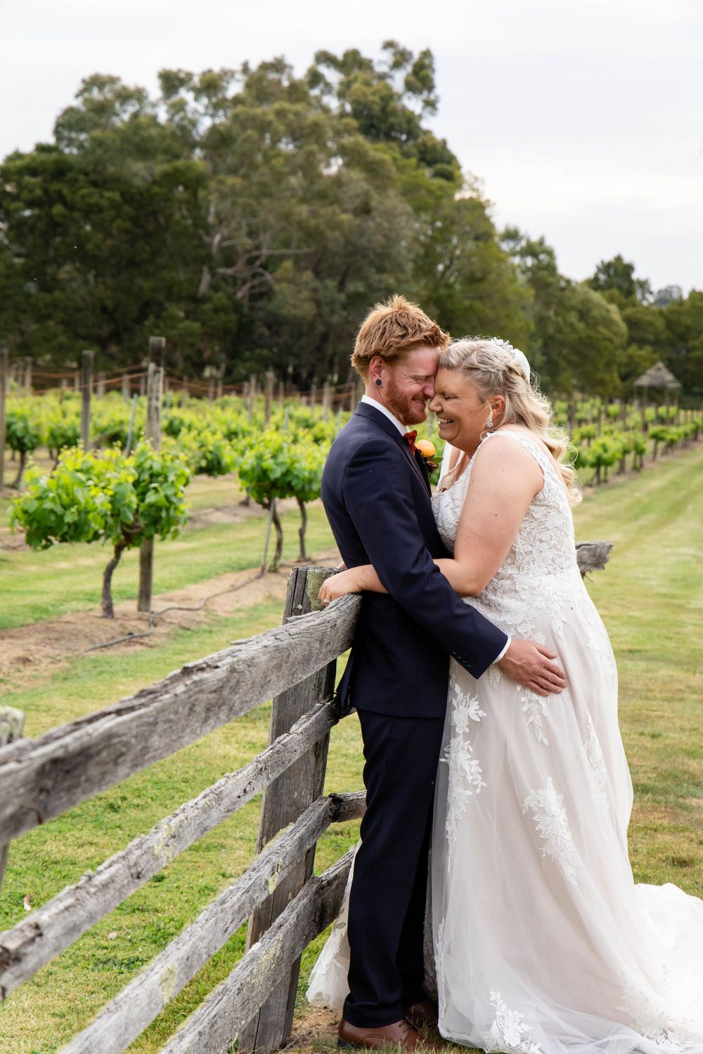 Newlyweds share an intimate moment at a rustic vineyard fence surrounded by grapevines and lush greenery.
