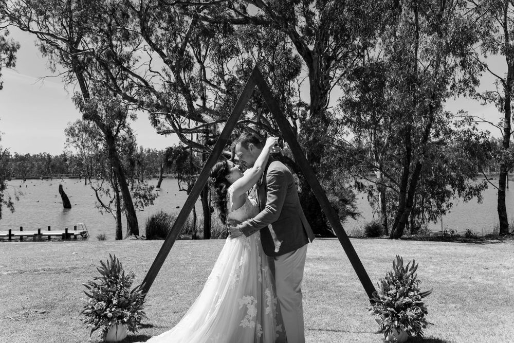 A couple embraces in wedding attire by a lake under trees with decorative plants in the foreground, captured in black and white.