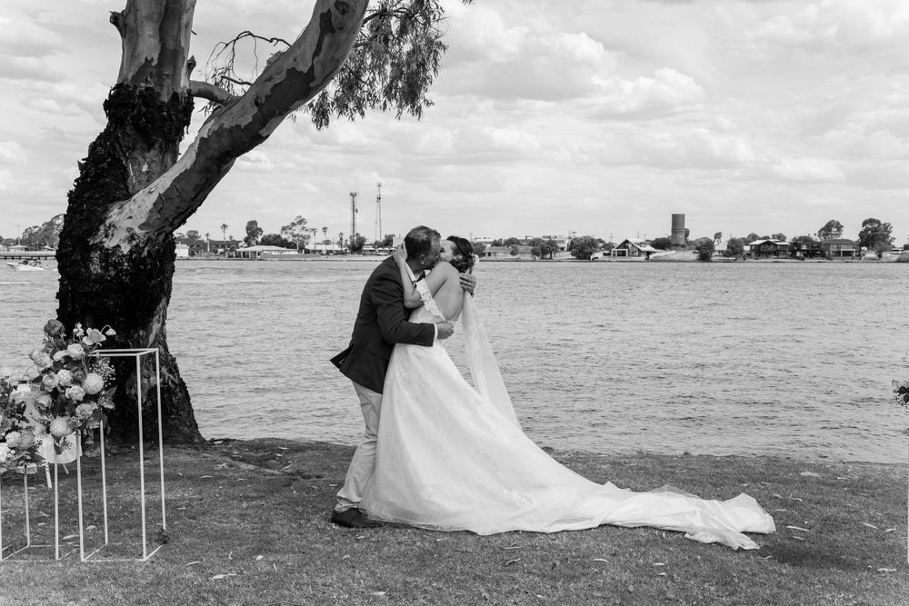 A couple in wedding attire embrace by a waterfront with a decorative tree and floral stands against a cityscape backdrop.