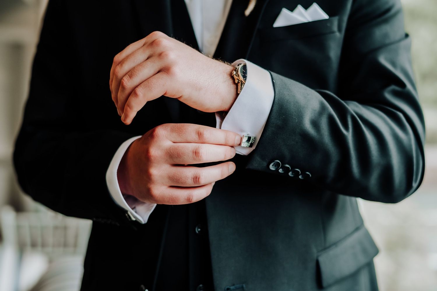 Close-up shot of hands adjusting a silver cufflink on a dark suit sleeve with crisp white shirt cuff.