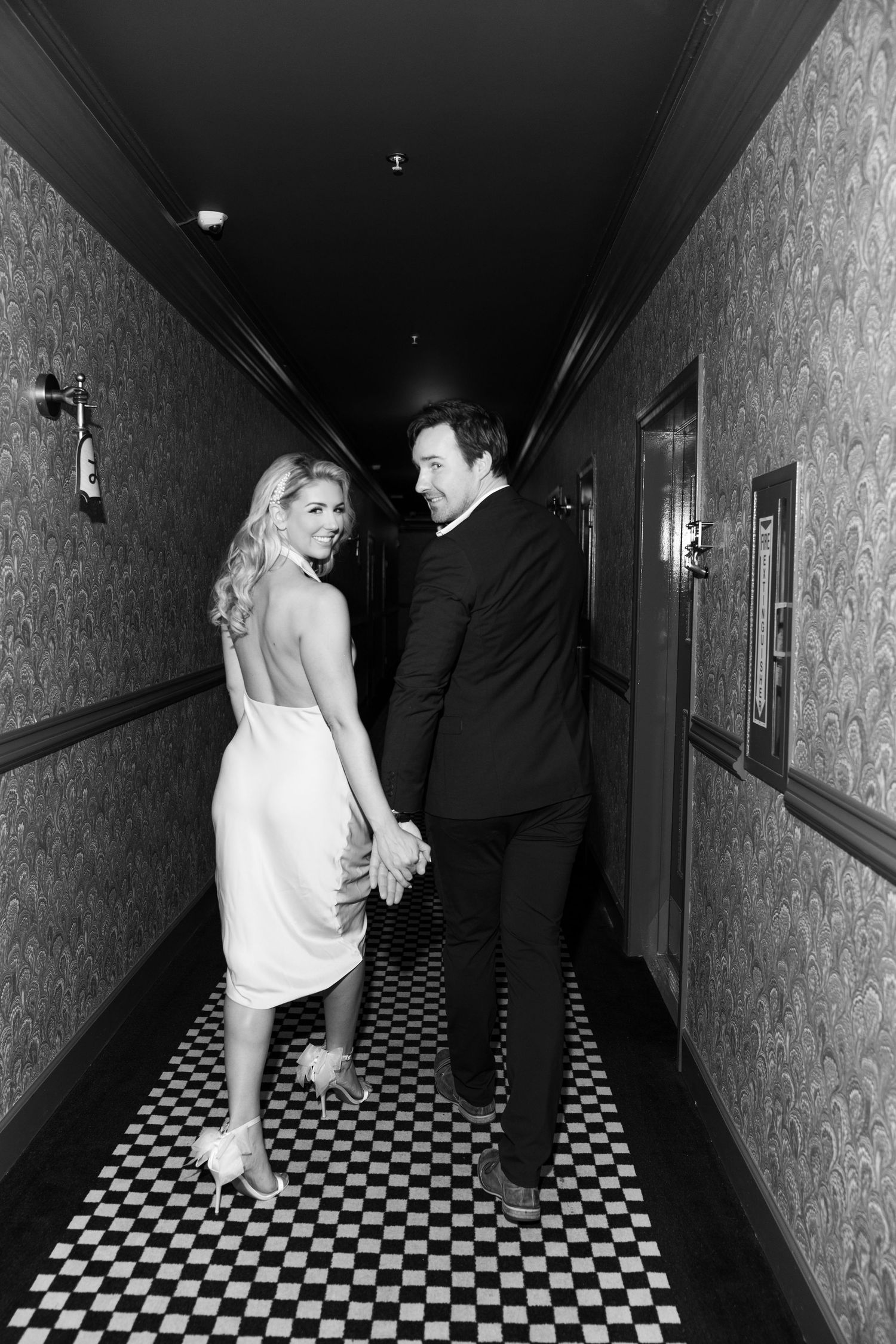 A couple walks down a vintage hallway with black and white checkered floors and floral wallpaper in classic evening attire.