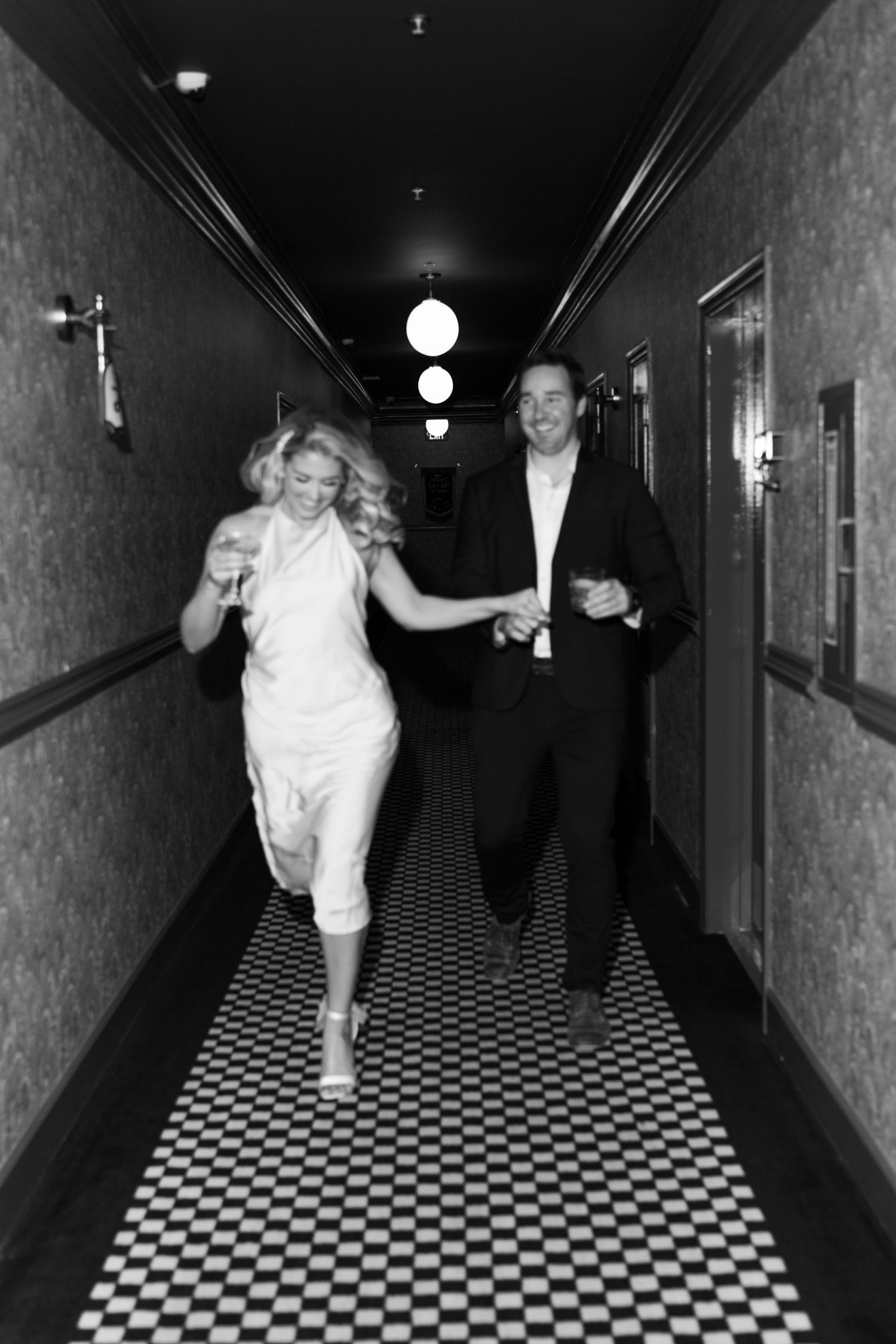 A couple runs joyfully down a vintage hallway in black and white photography. Engagement Session at The Lafayette Hotel in San Diego. JPPC