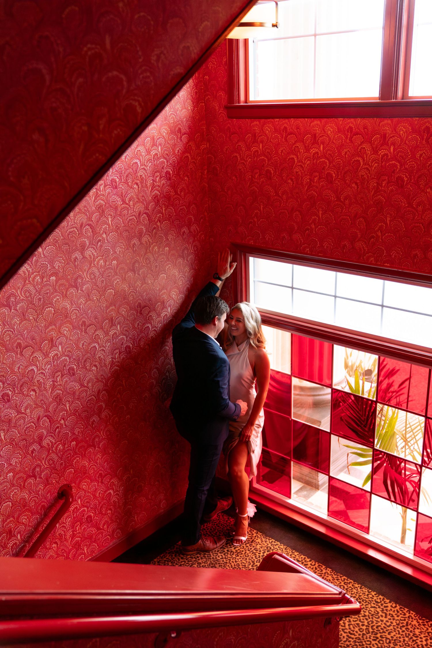 Couple standing by a stained glass window in a red stairwell with dramatic lighting. Engagement Session at The Lafayette Hotel in San Diego.