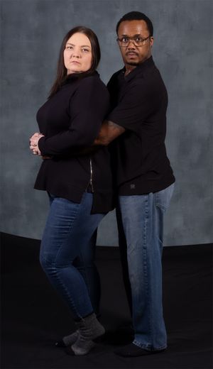 Couple in matching black tops and jeans pose back-to-back in studio setting.