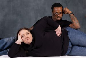 A playful studio portrait of a couple in black shirts posing casually against a gray backdrop.