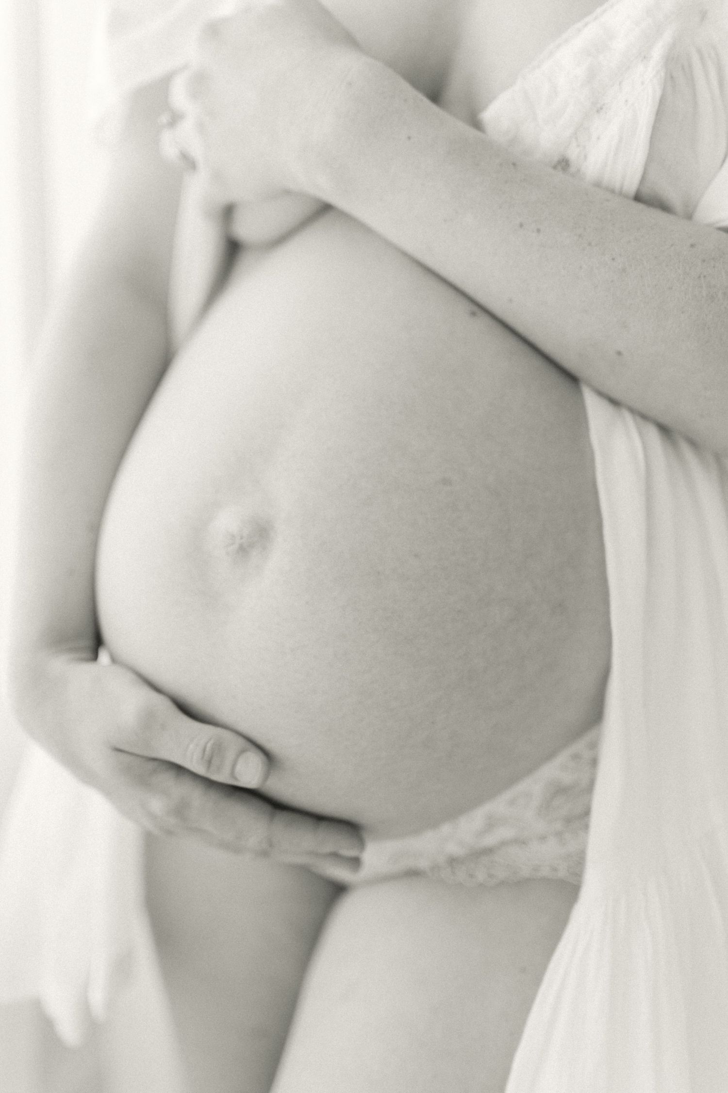 Artistic black and white maternity photo showing hands gently cradling a pregnant belly in soft lighting.