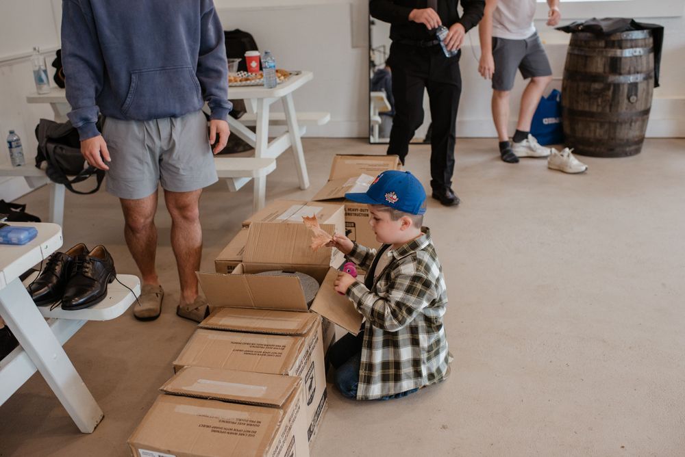 Someone wearing a plaid shirt sits on a floor against cardboard boxes while others stand in a room with white benches.