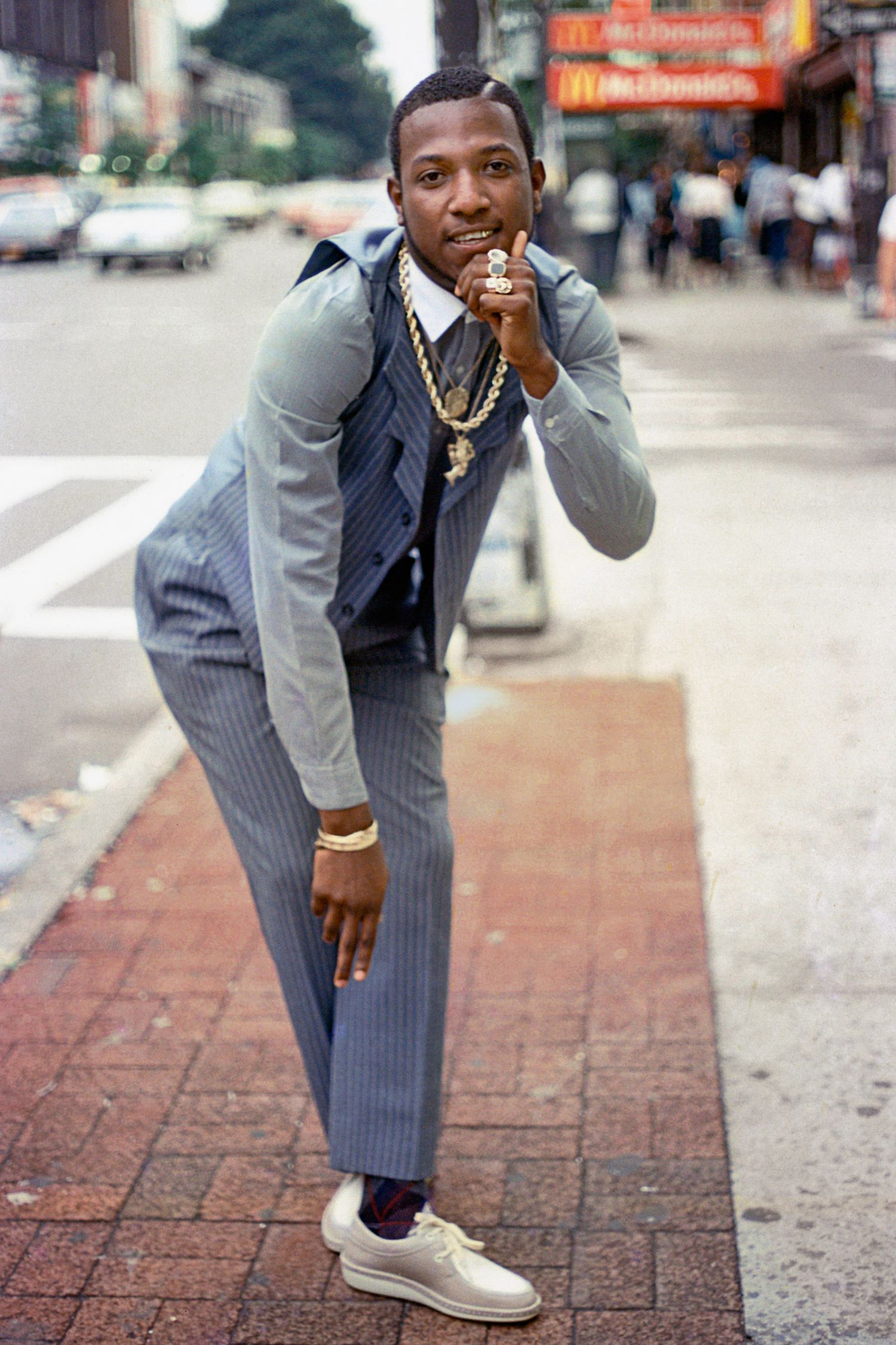 Stylishly dressed person in gray suit striking playful pose on city sidewalk.