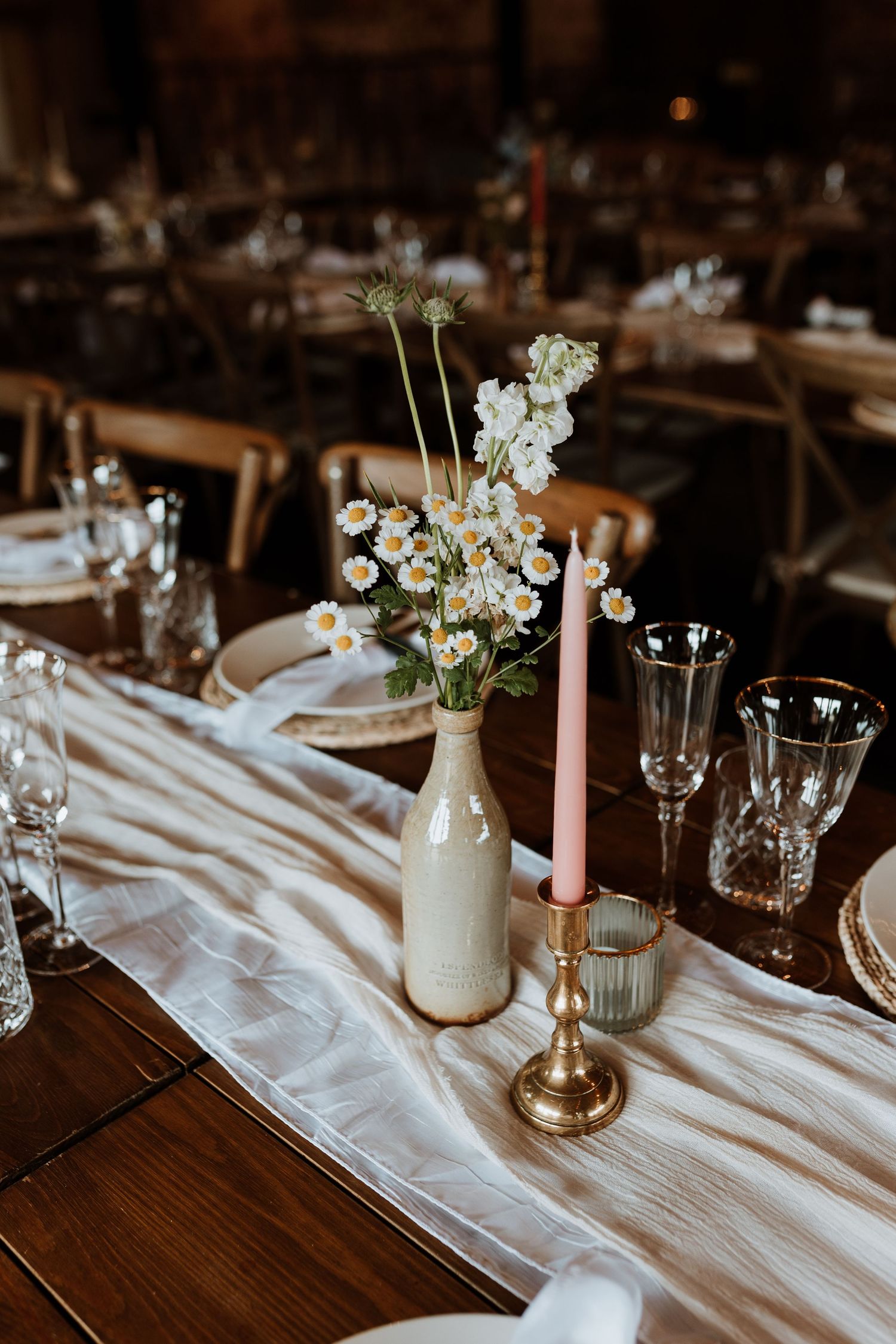 Rustic wedding table setting with white floral centerpiece in milk bottle vase, pink candle, and crystal glassware on linen runner.