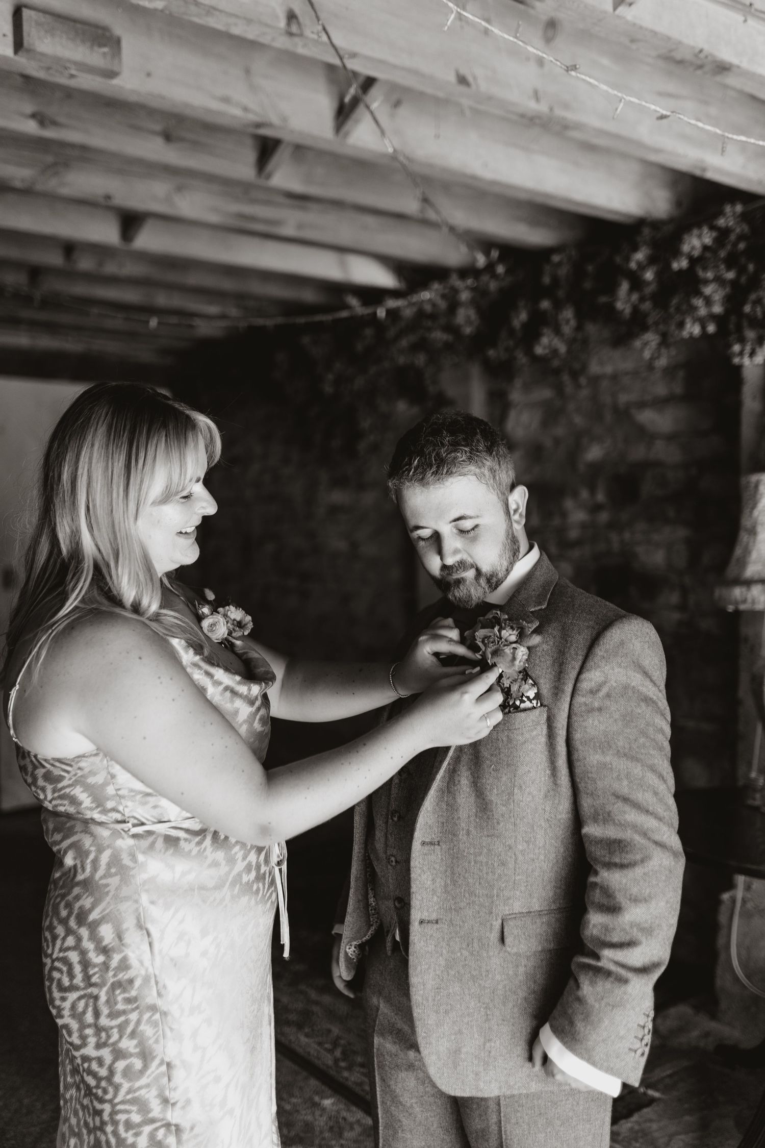 A tender moment during wedding preparations as a boutonniere is pinned onto a gray suit jacket in black and white at Lyde Court.