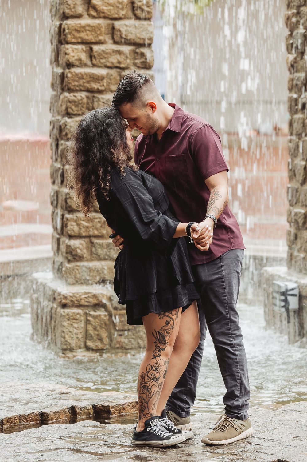 A romantic couple embracing by a water fountain wearing casual outfits.