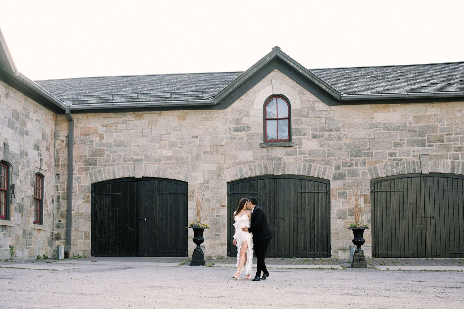 A couple embraces in front of rustic stone stables with dark green wooden doors and traditional architecture.