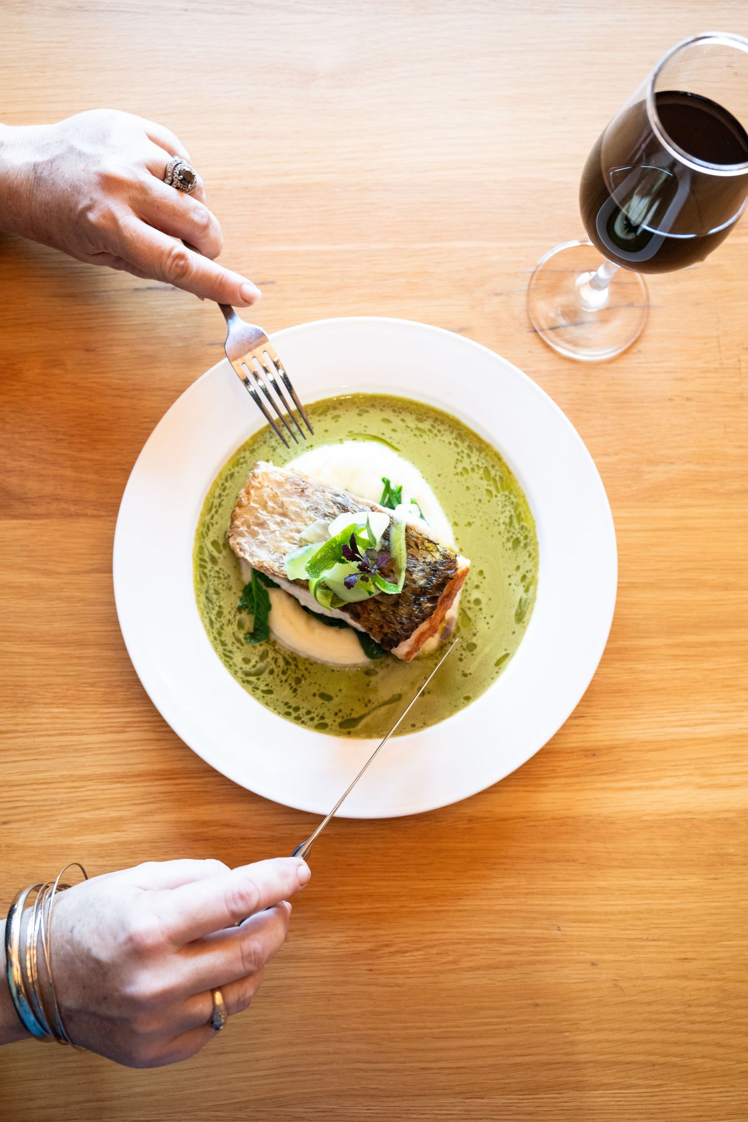 Hands holding utensils over a plated fish dish with vibrant green sauce on a white plate against a wooden table surface.