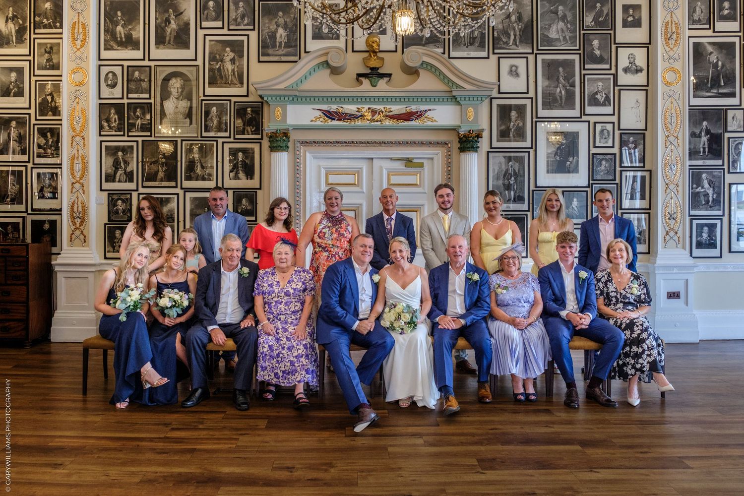 Wedding guests sit in formal attire against a wall decorated with vintage black and white photographs and portraits.