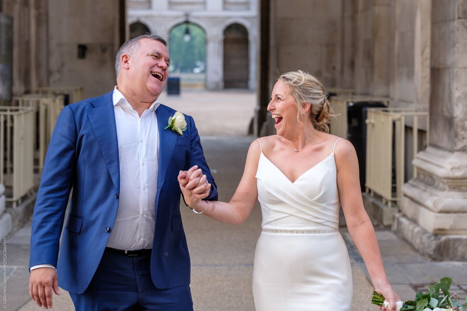 Newlywed couple laugh together while walking through historic stone archway passage in matching blue and white attire.
