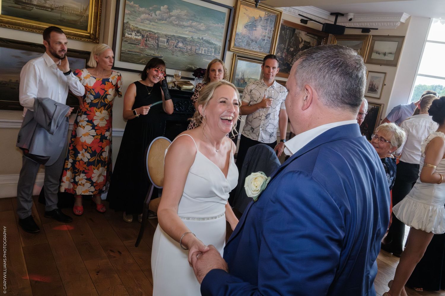 Wedding guests dance together on a wooden dance floor in a room decorated with framed artwork.