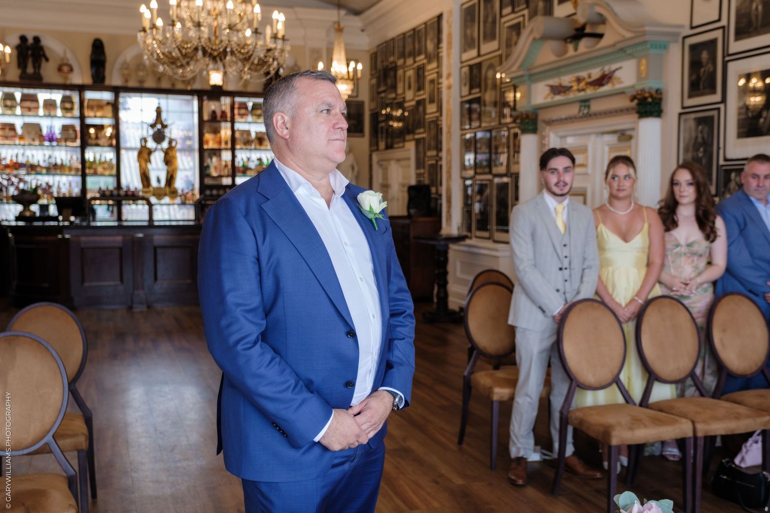 An elegant indoor wedding ceremony setting with vintage chairs, chandeliers and a bar area with glass displays.