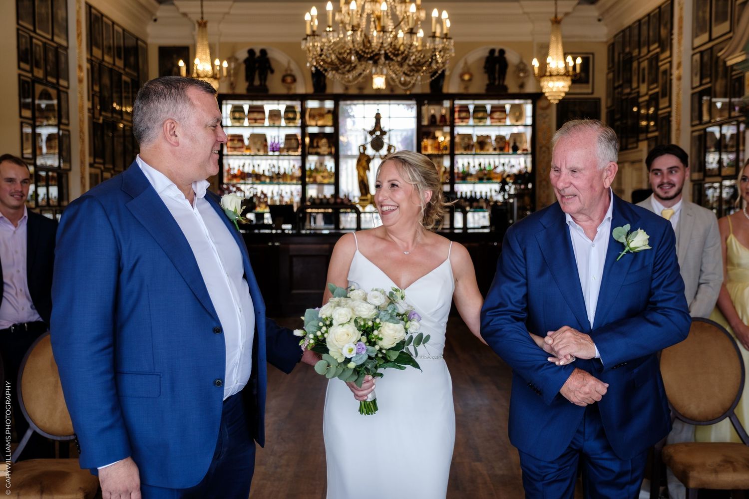 Three people standing in an elegant restaurant bar area with chandeliers, with someone in a white wedding dress holding flowers.