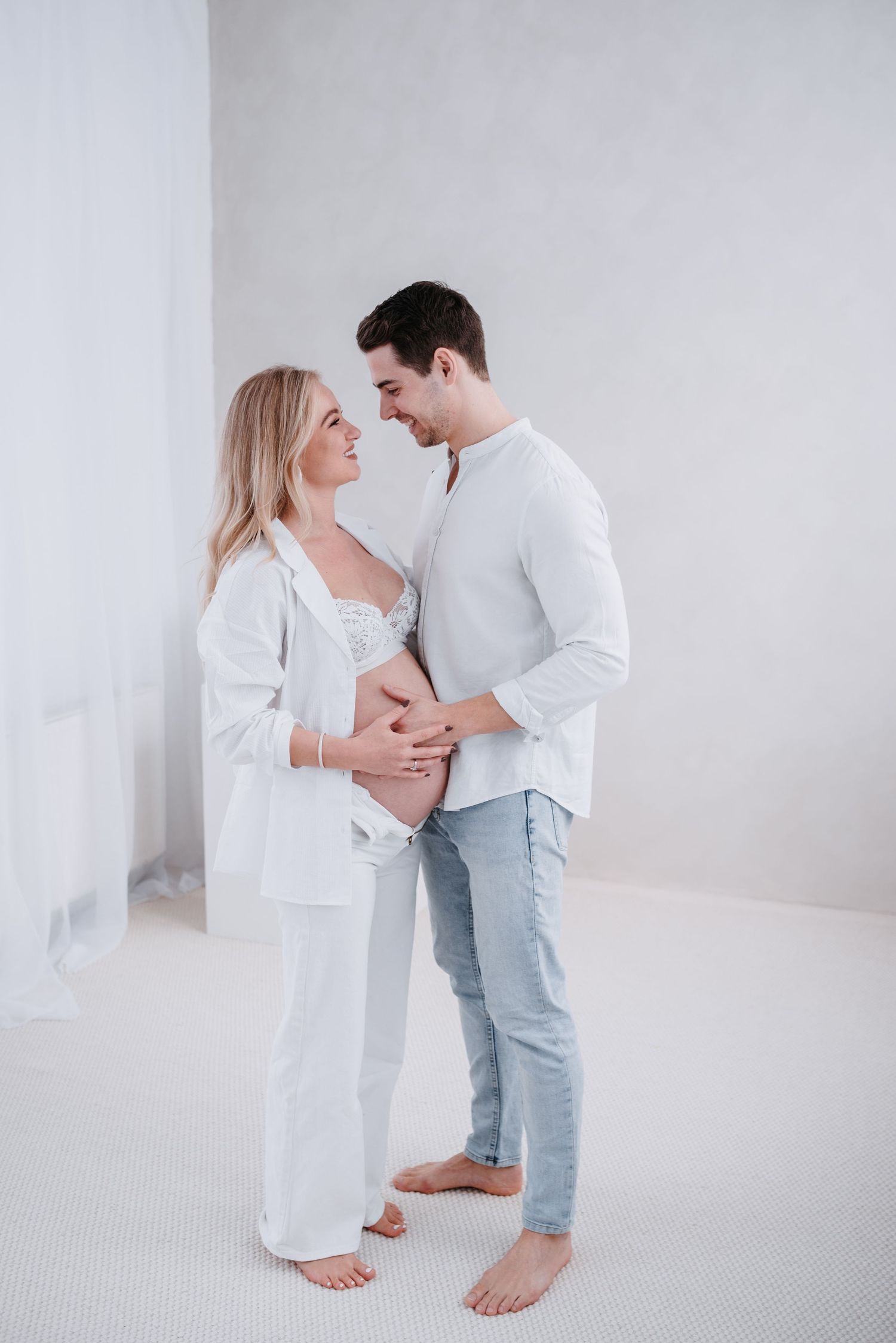 A couple in white clothing share an intimate moment while expecting a baby in a bright minimalist setting.