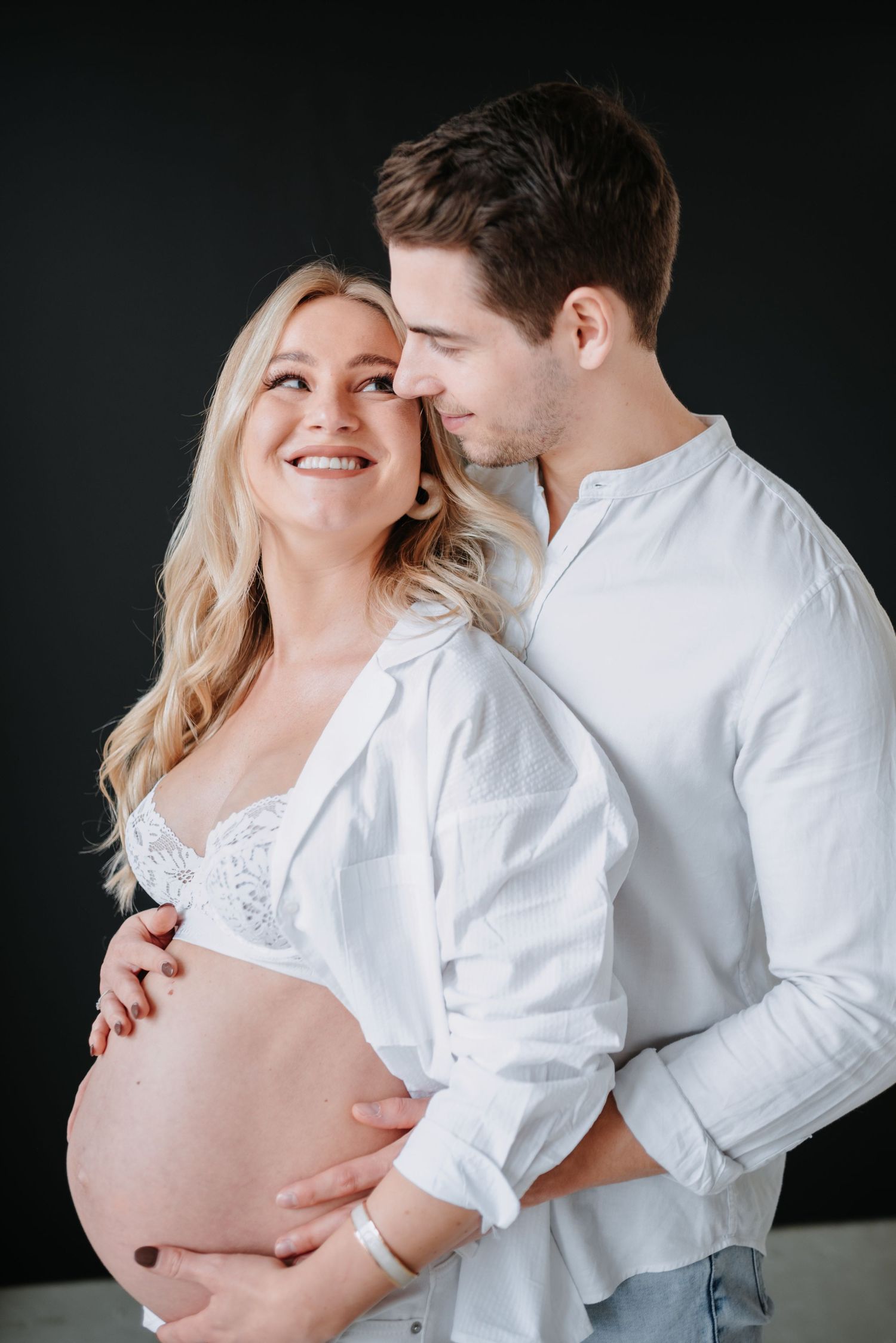 Intimate maternity photo of couple in white shirts sharing a tender moment against dark background.