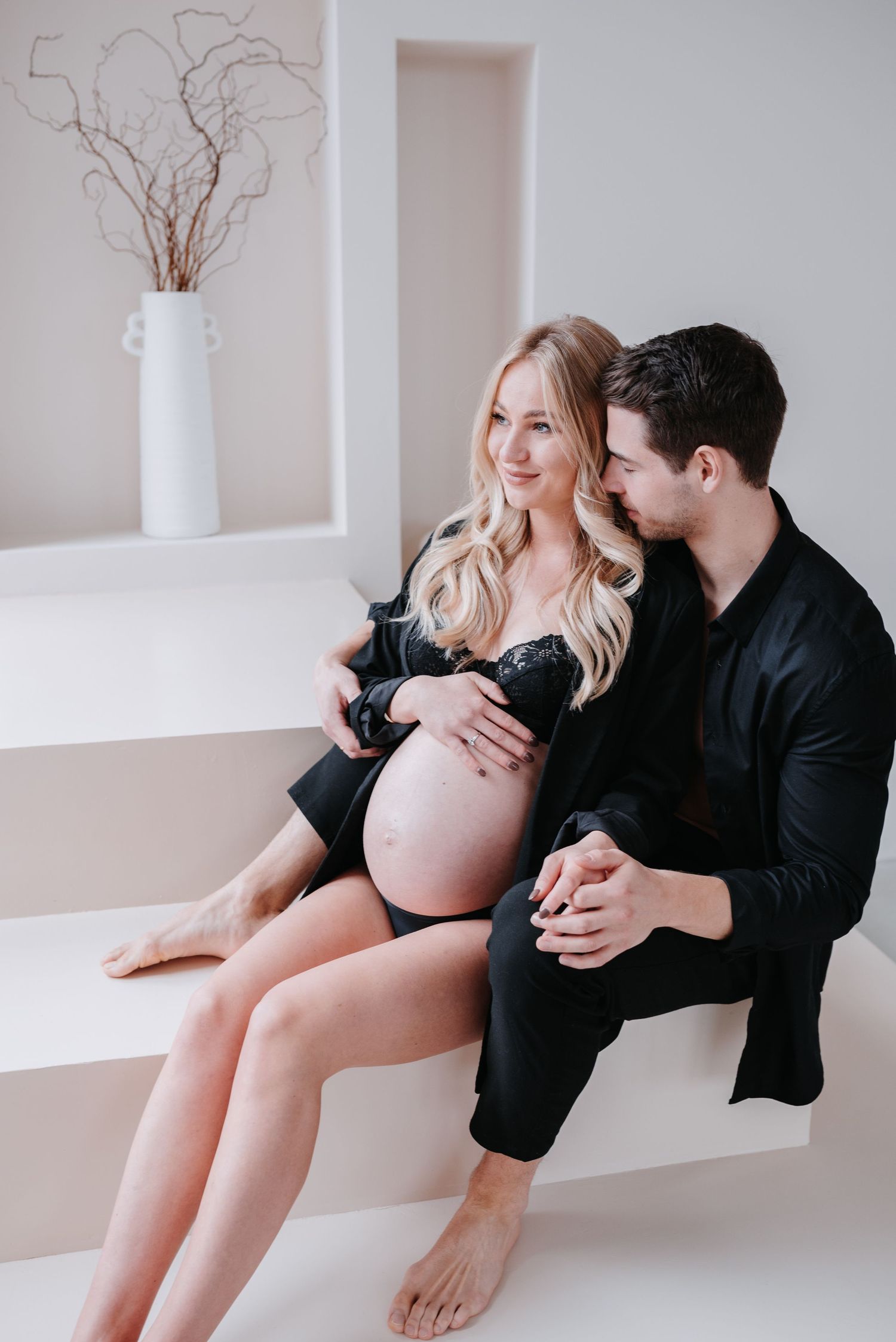 A maternity photoshoot series shows a couple sharing tender moments together against a minimalist white wall backdrop.
