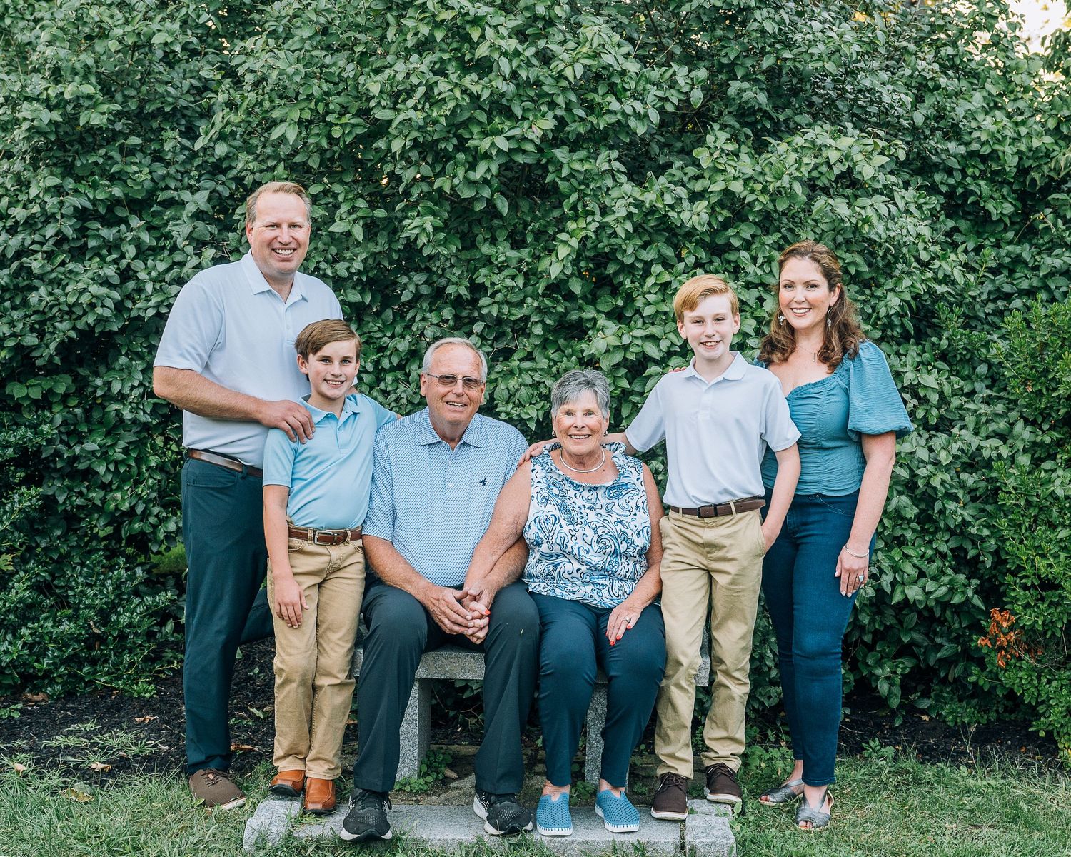 Multi-generational group in coordinated blue and khaki outfits poses outdoors against green foliage background.