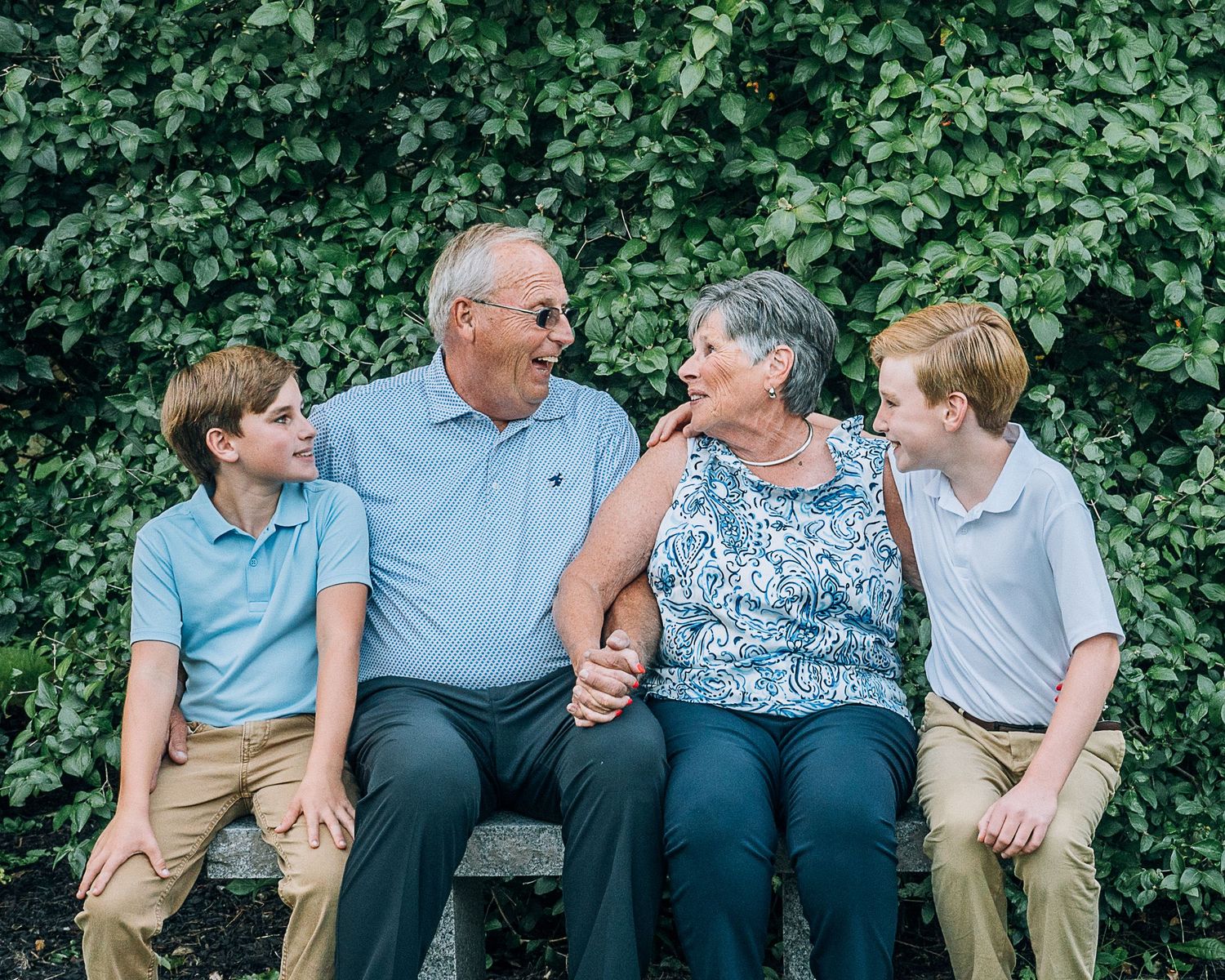 Multi-generational family sitting together on bench against green leafy backdrop, sharing loving smiles and laughter.
