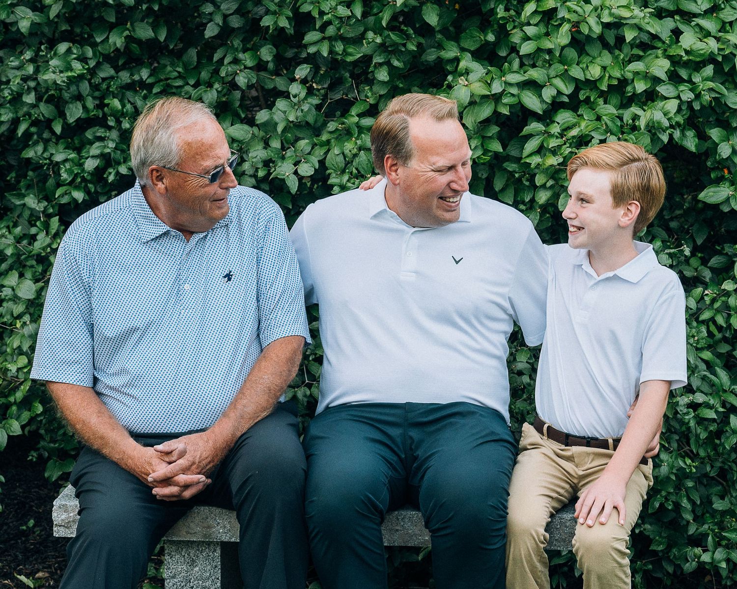 Three generations in coordinating polo shirts share a candid moment together while sitting outdoors against green foliage.