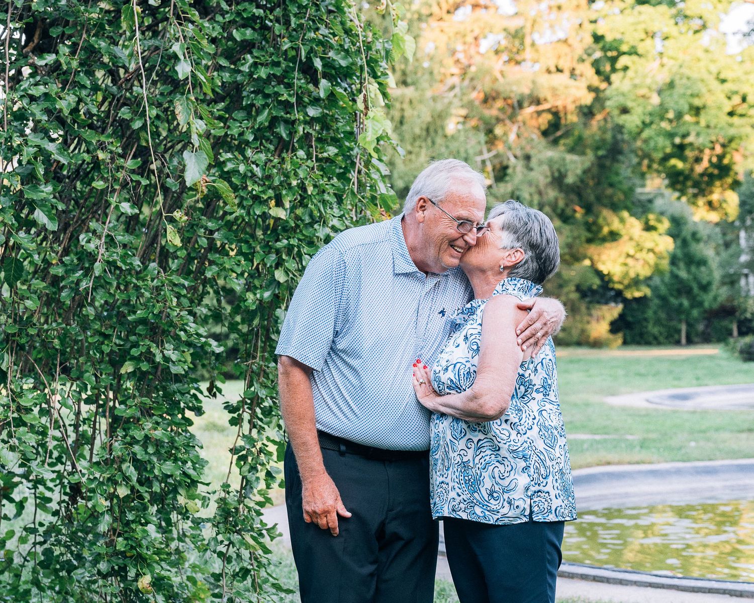 Elderly couple sharing tender moment in garden setting with green foliage and autumn colors in background.