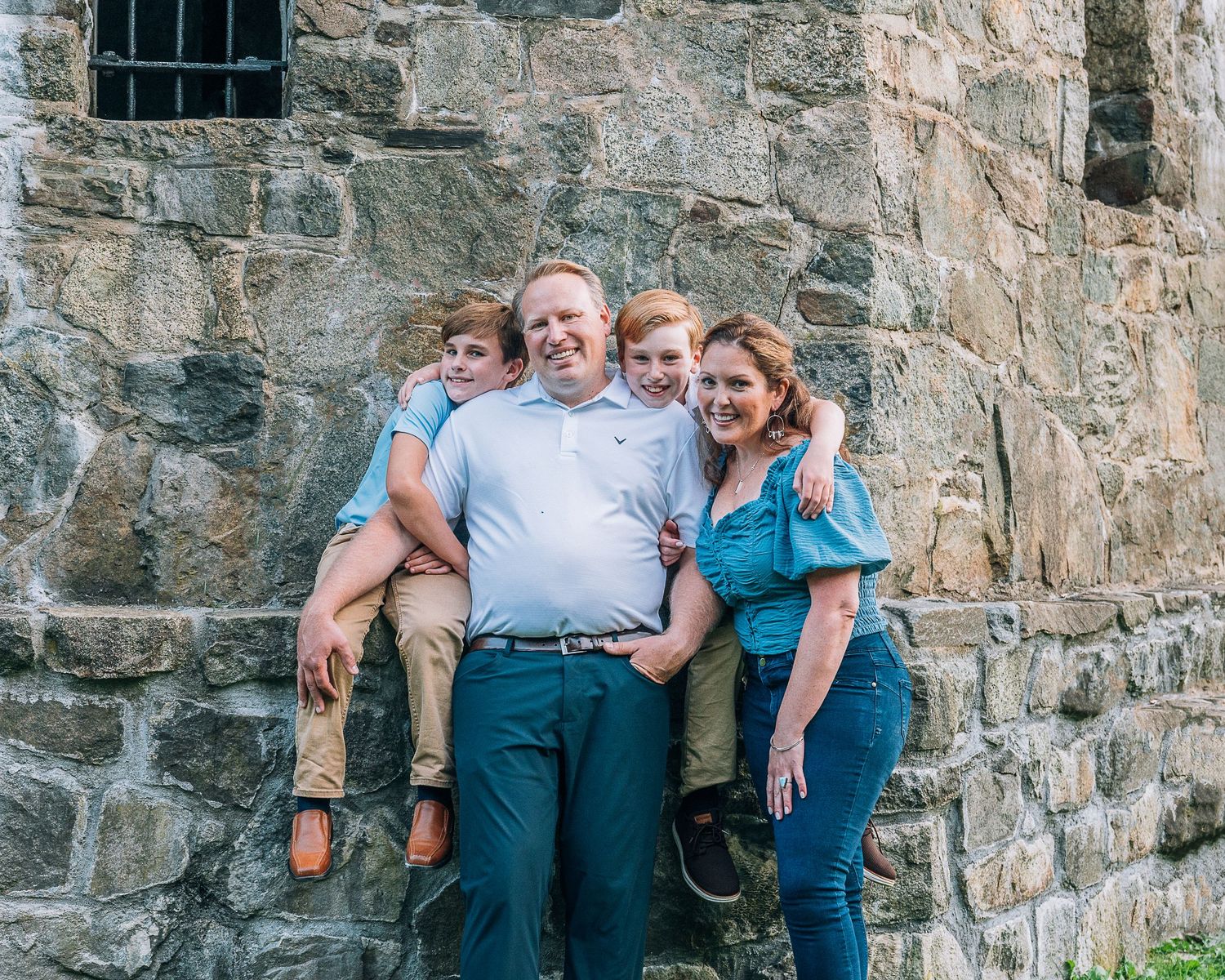 A group of three people pose together against a rustic stone wall on a sunny day outdoors.