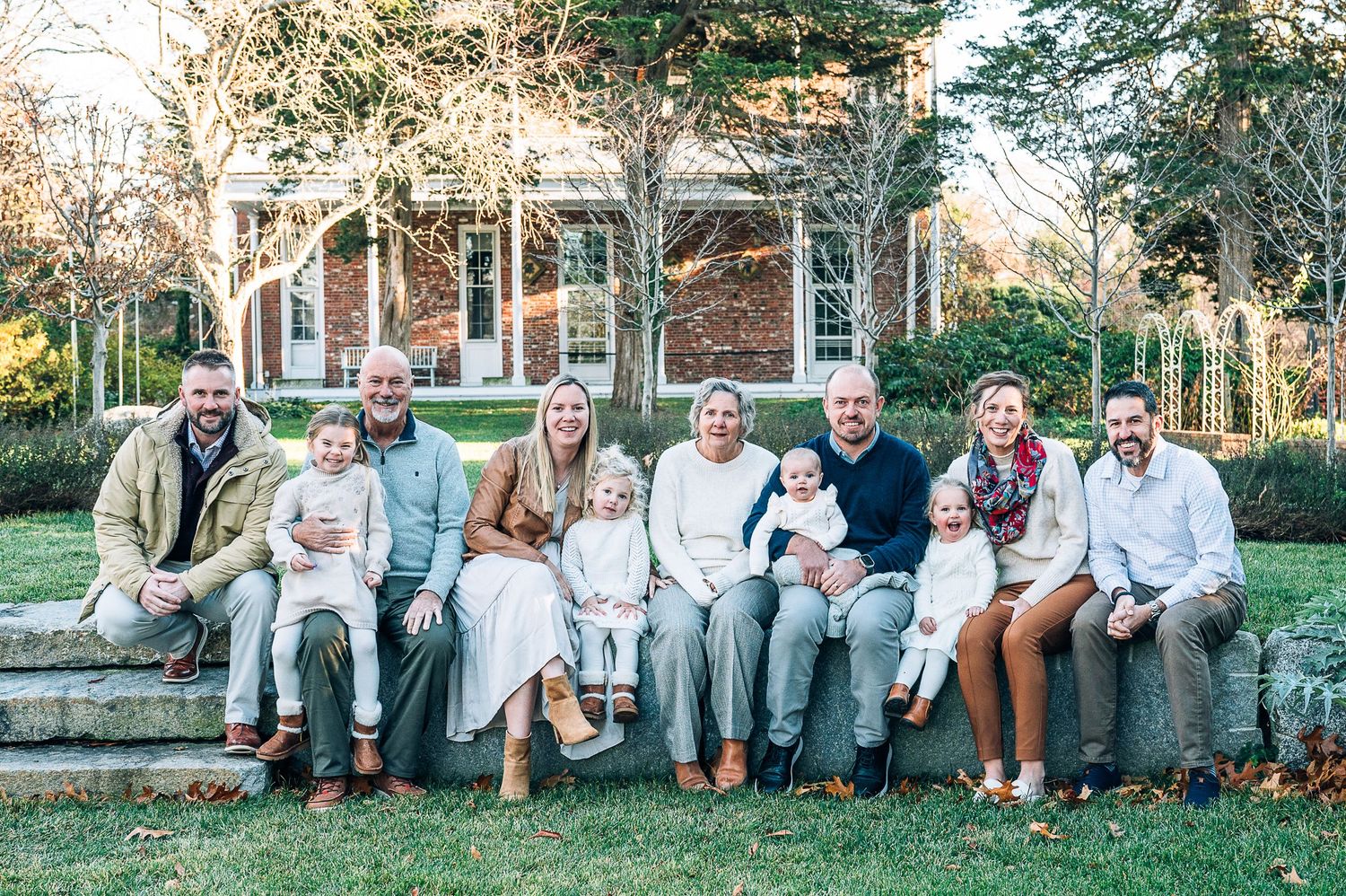 Extended family group sits together outdoors along stone wall in coordinated neutral clothing for fall photo session.