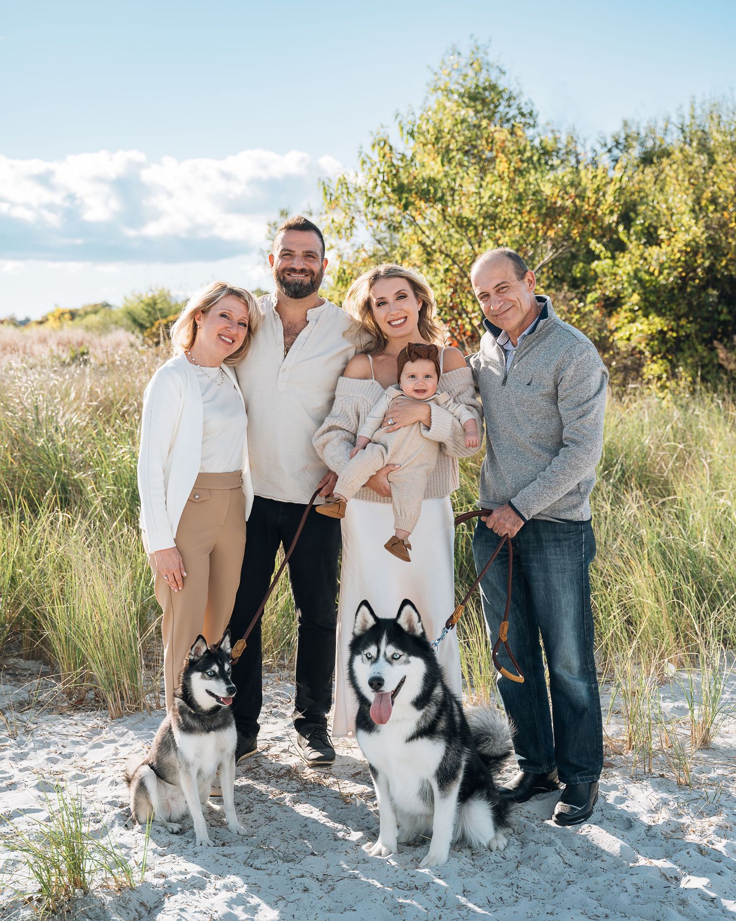 A family and their two Siberian Husky dogs pose together on sandy terrain with greenery in the background.