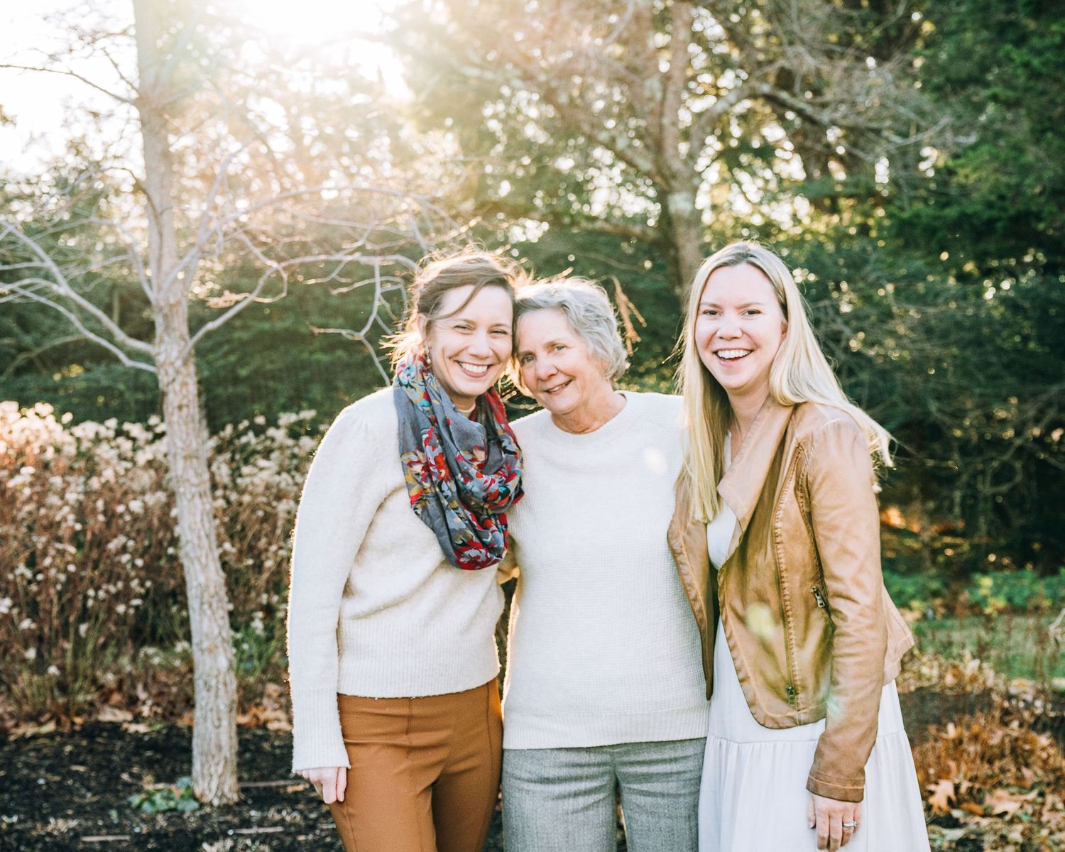 Three people pose for a casual portrait outdoors with sunlight filtering through autumn trees behind them.