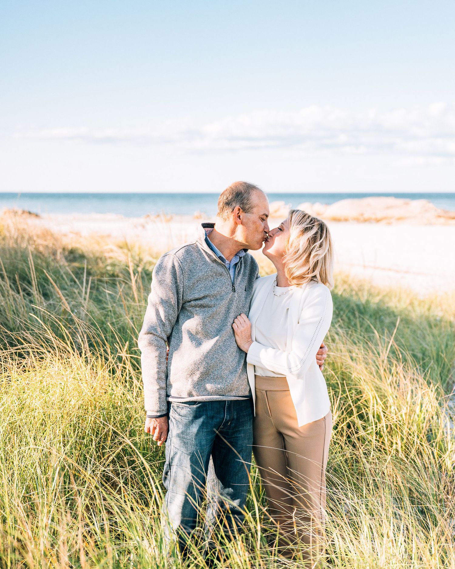 A romantic couple shares a tender moment among beach grass with the ocean and sky in the background on a sunny day.