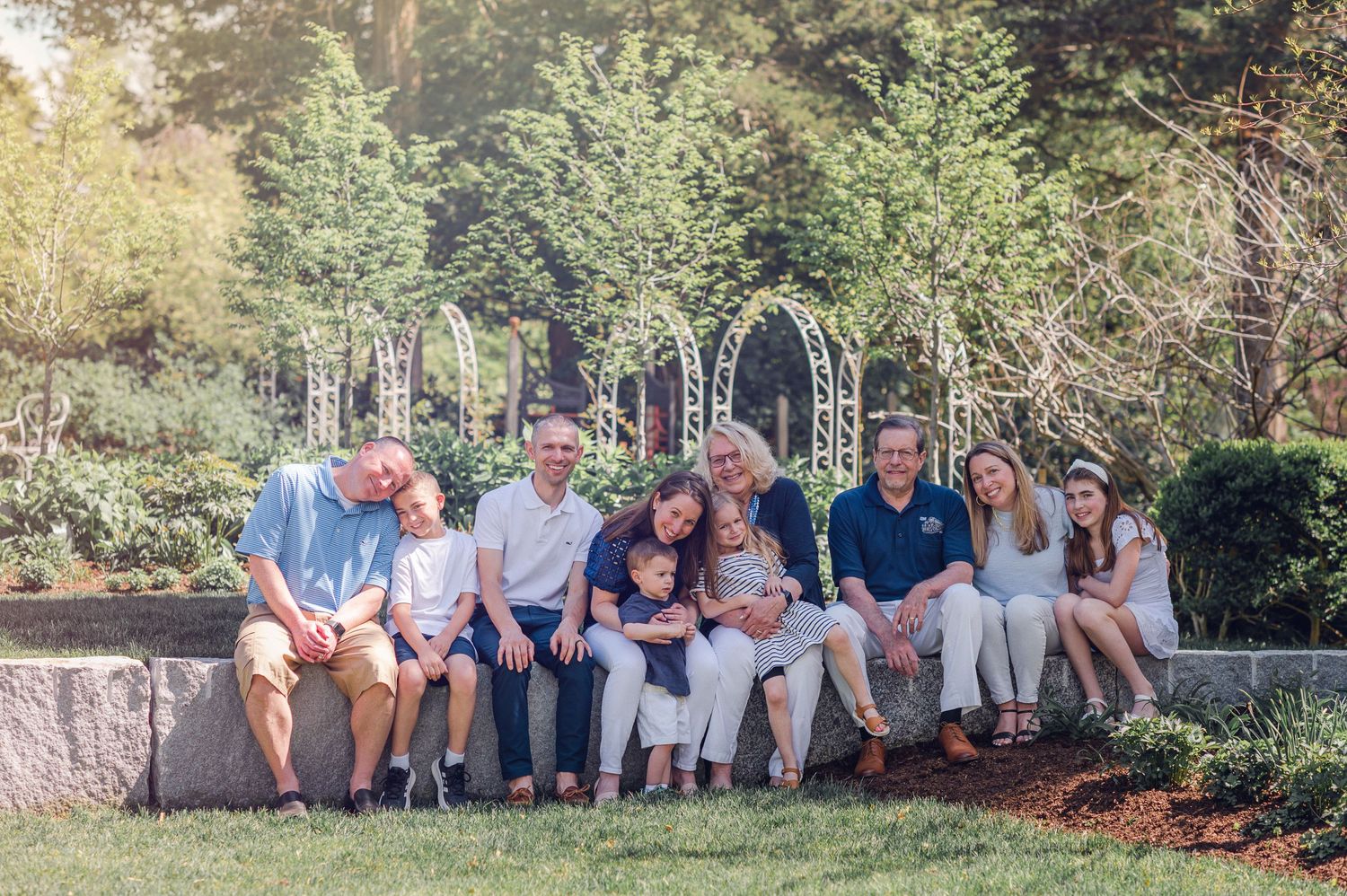 Group sitting together on stone wall in garden setting with decorative arches and greenery in background.