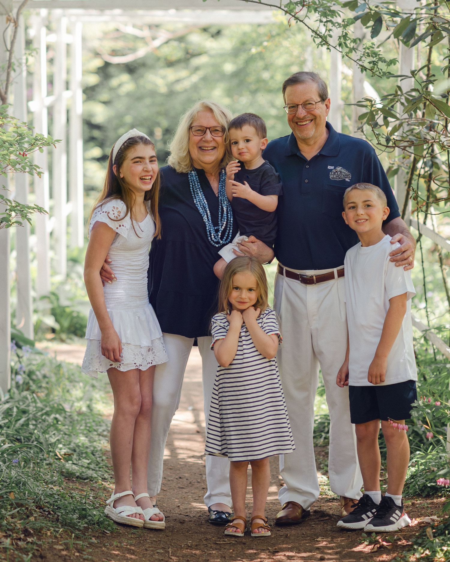 A multigenerational family poses together outdoors on a wooded path in coordinated light colored clothing.