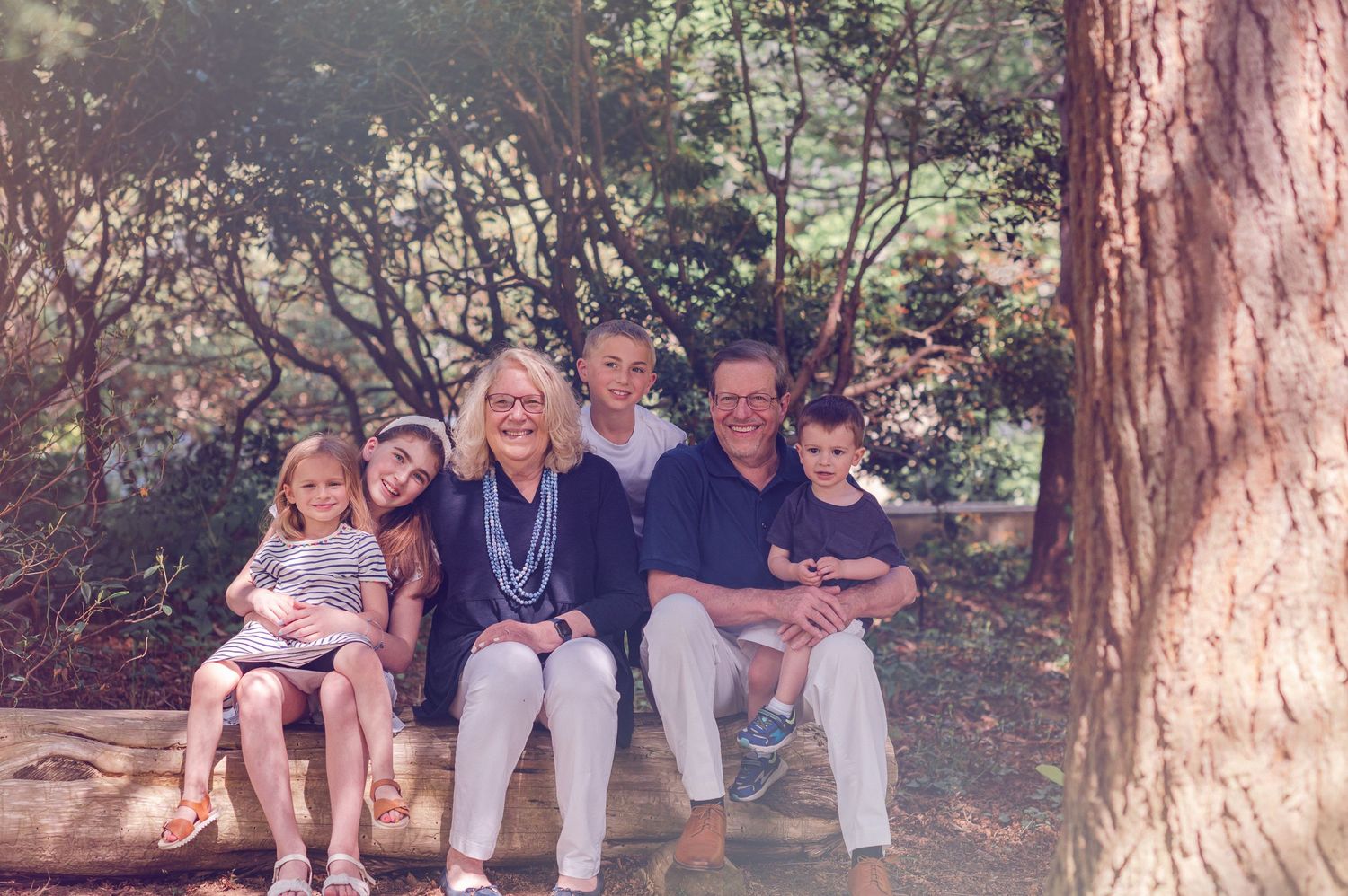 Family group sits together on outdoor wooden bench near tall tree trunk in natural setting.