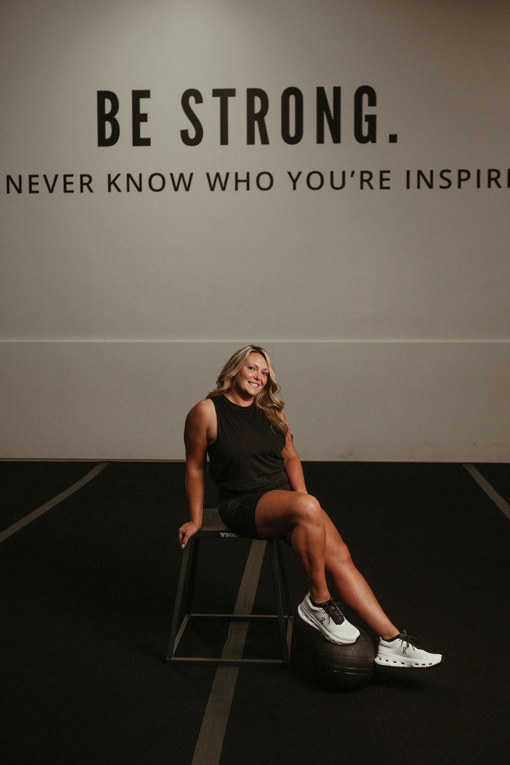 Athletic woman sits on modern chair beneath motivational wall text reading 'BE STRONG' in dark minimalist gym setting.