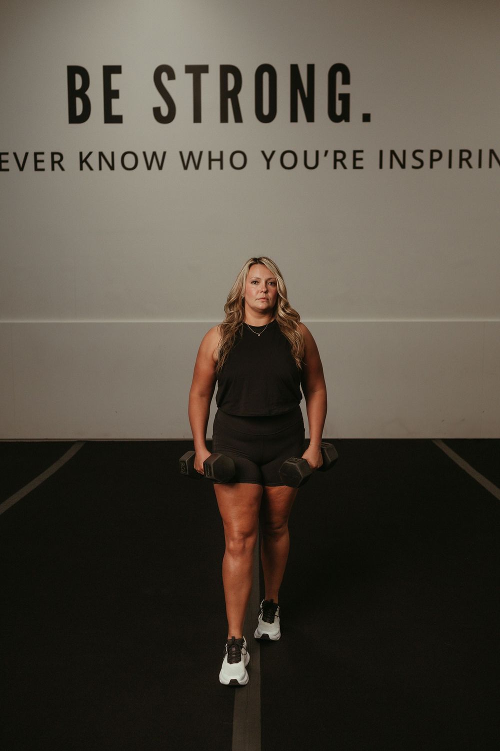 Athletic woman in dark workout attire stands confidently beneath motivational wall text reading BE STRONG.