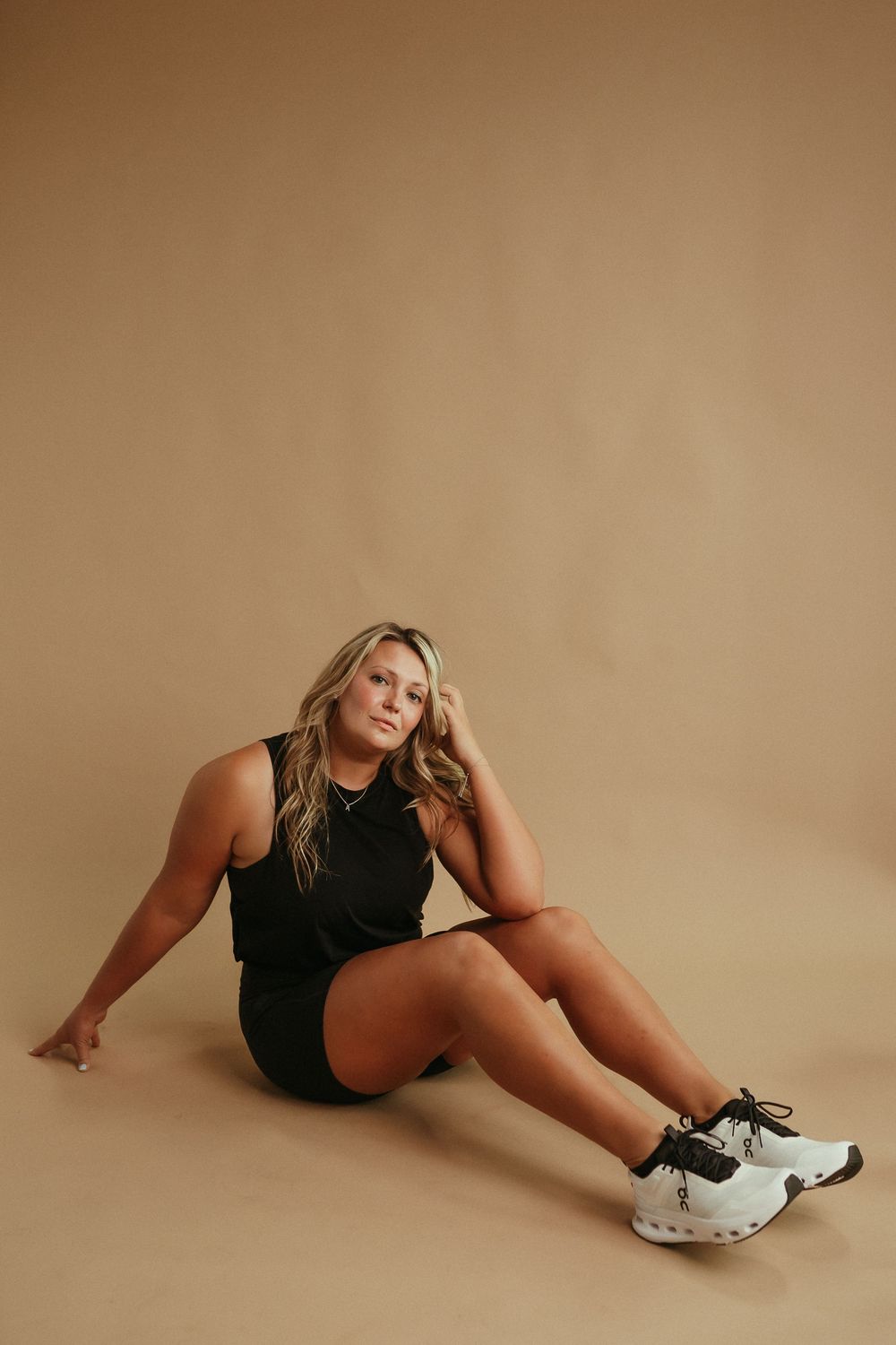Athletic model in black outfit and sneakers poses on brown background in relaxed seated position.