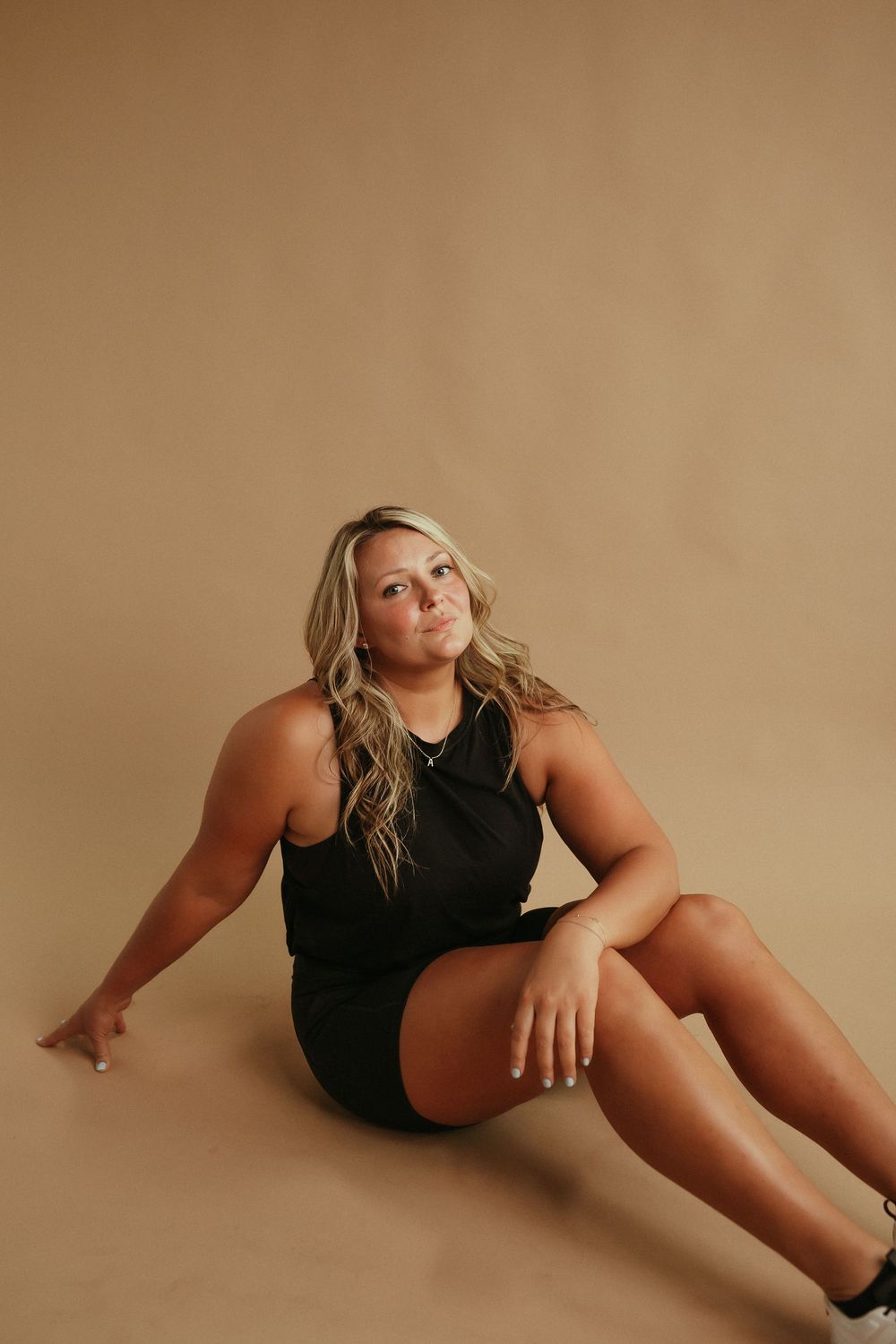 Athletic woman in black outfit poses gracefully against beige backdrop during fitness photoshoot.
